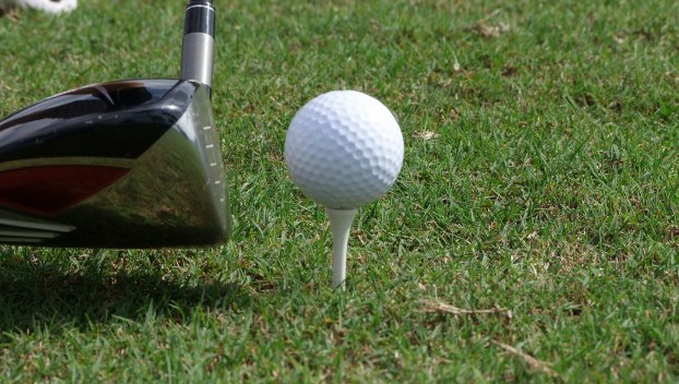 A golf ball sits on a tee in the grass with a driver lining up a shot to the left