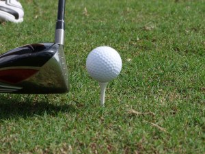 A golf ball sits on a tee in the grass with a driver lining up a shot to the left