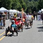 Cyclists and walkers stroll artist vendor lined street during the 2025 Troutdale Arts Festival on a sunny day.